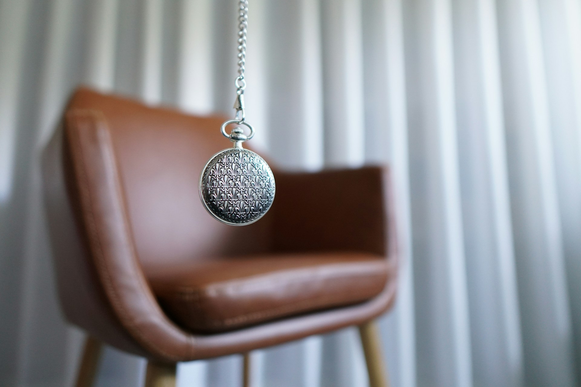 A silver pocket watch hanging in front of a brown leather chair.